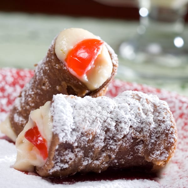 Two cannolis, one dusted with powdered sugar, filled with cream and candied cherry on a red plate.